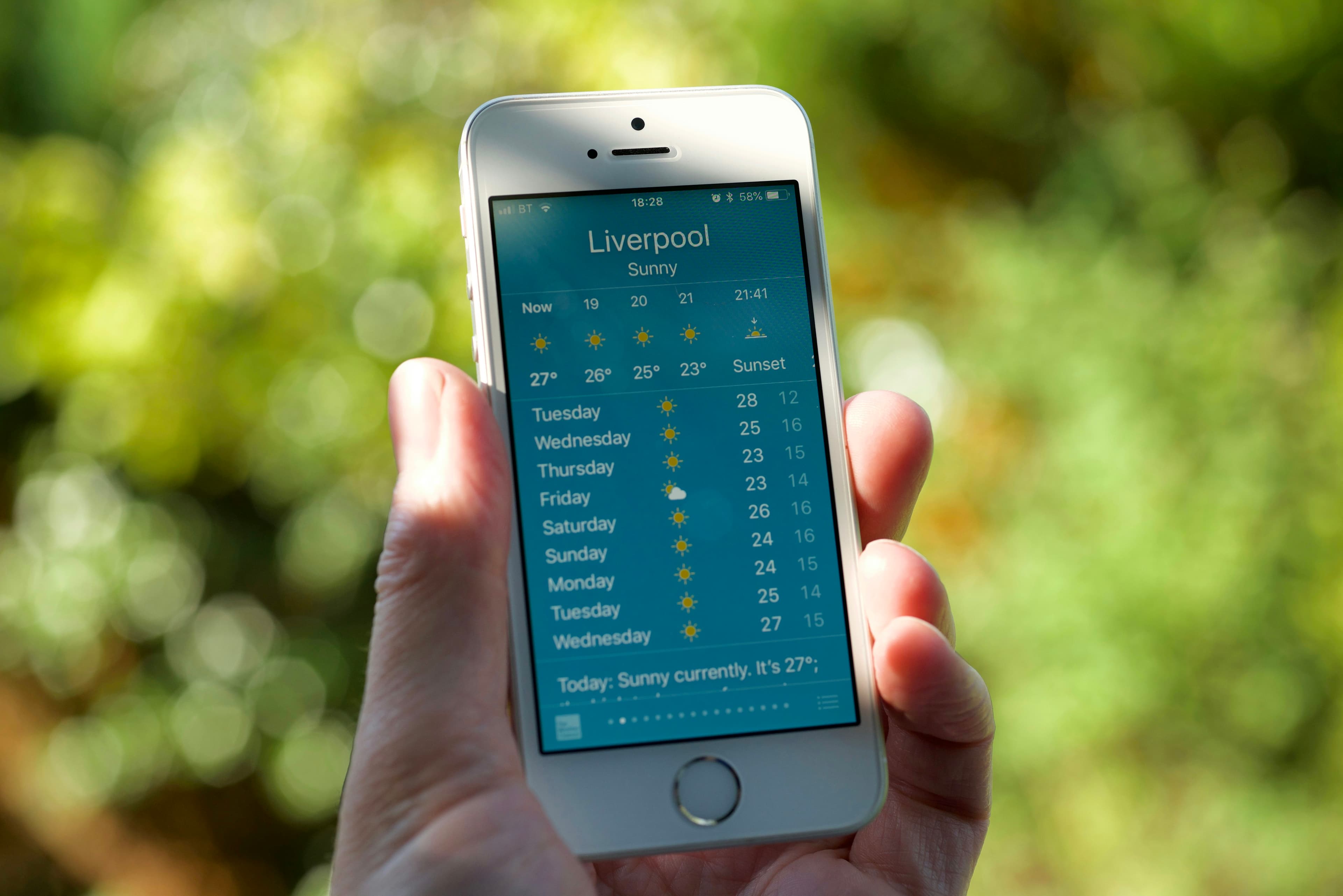 A close-up shot of a hand holding a smartphone displaying a sunny weather forecast for Liverpool. The vibrant teal and yellow color scheme of the app contrasts beautifully with the blurred green background, creating a calming and optimistic mood. The sha…