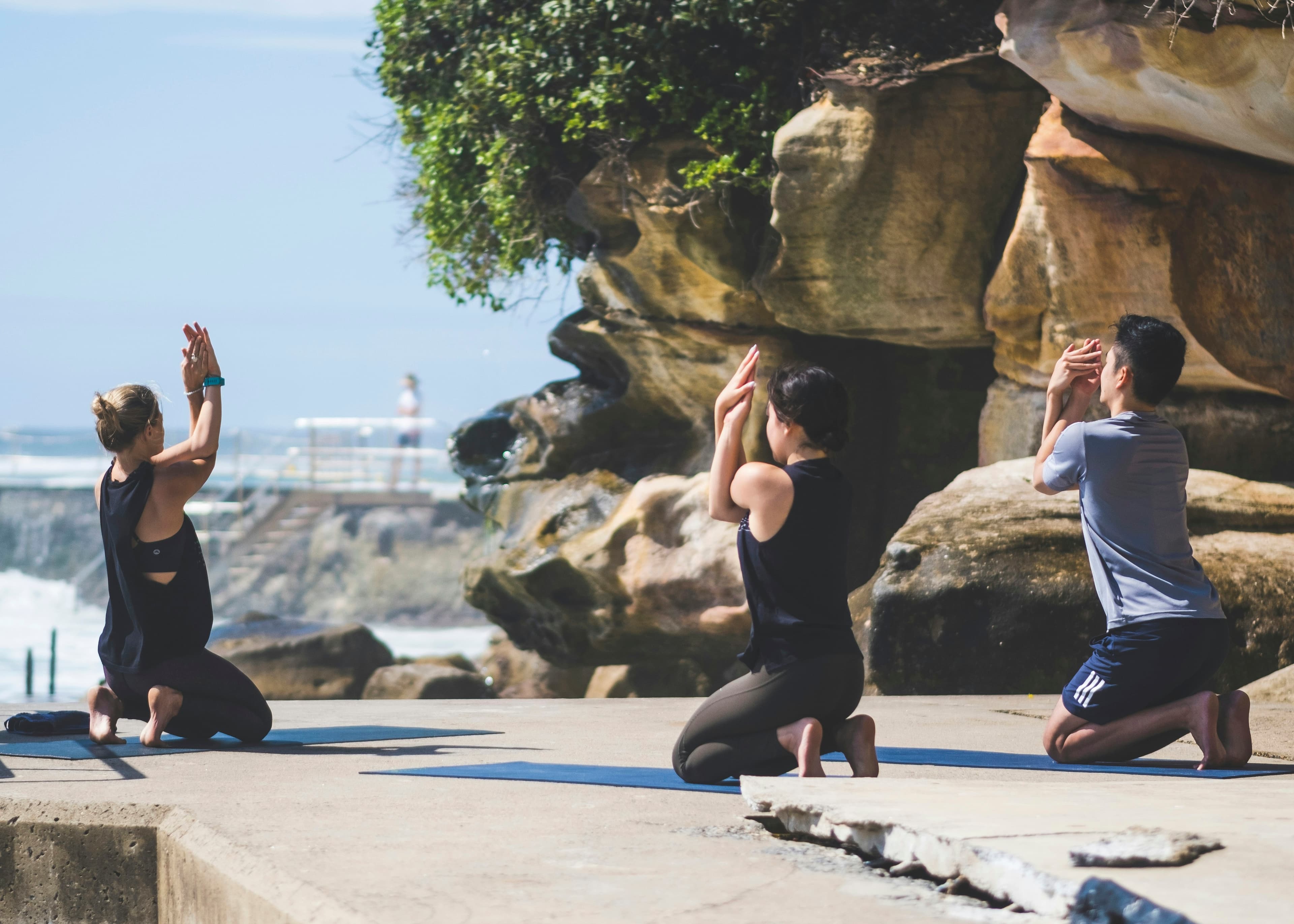A small group does yoga on the beach on a sunny day, near the water and beautiful rocks.