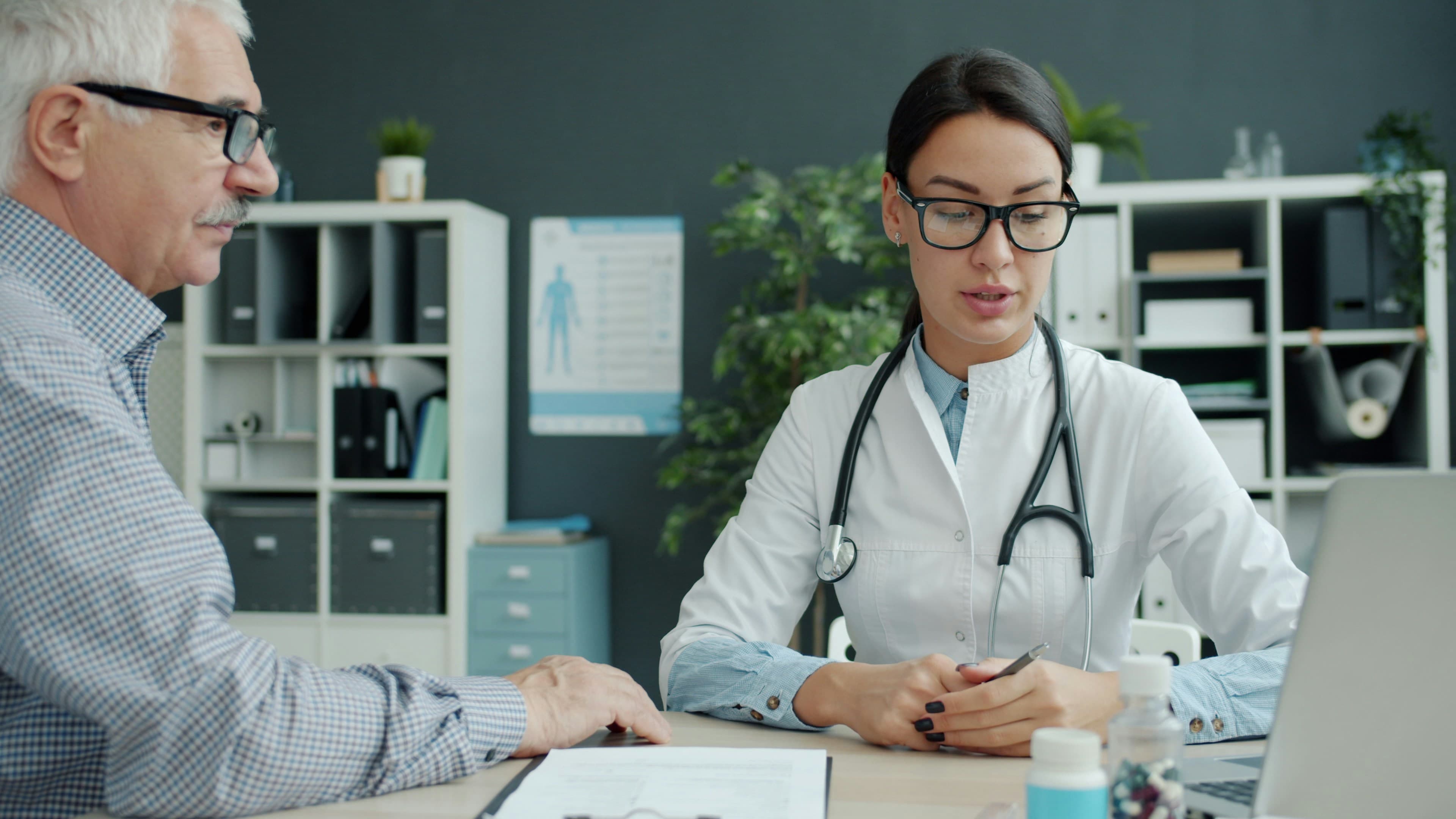 Caring female doctor is talking to elderly man patient holding hand expressing care in clinic. Healthcare, people and communication concept.